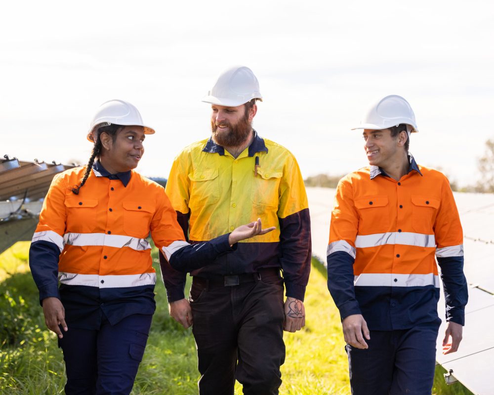 Three Technicians Surrounded By Solar Panels On Solar Farm in Australia. Mutiracial Mix Of One Caucasian And Two Aboriginal Australians